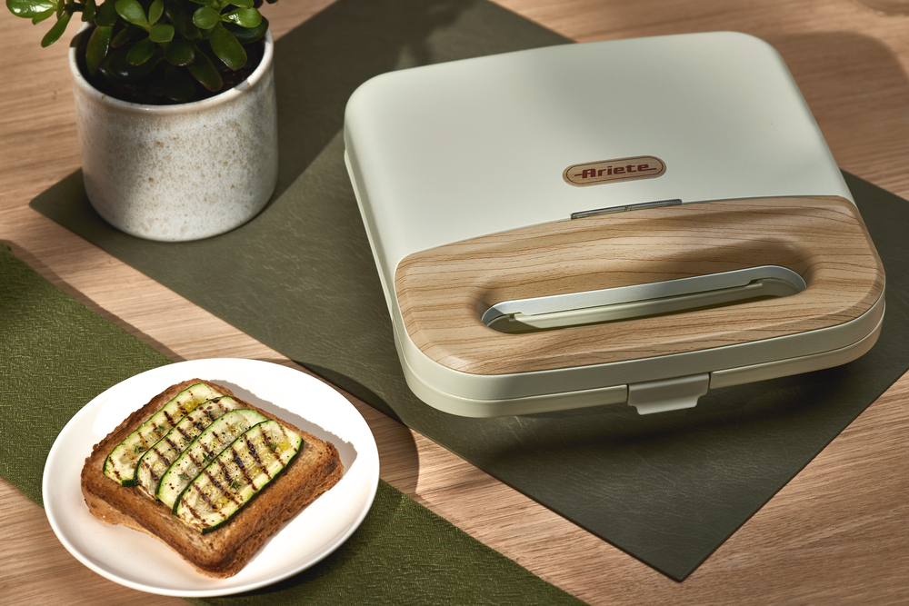 Тостер за сандвичи „Breakfast Wood“ с дървен акцент на дръжката стои на маса до саксийно растение и чиния с филия препечен хляб, гарнирана с авокадо и подправки.
