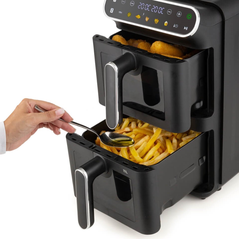 Ръка с лъжица се промъква в долното чекмедже на Air Fryer Vertical Duo – фритюрник с горещ въздух с две кошници, 11 л, пълно с картофи фри; горното чекмедже съдържа панирани храни. Наблизо робот-прахосмукачка работи тихо, осигурявайки чистота във всяка част на кухнята.