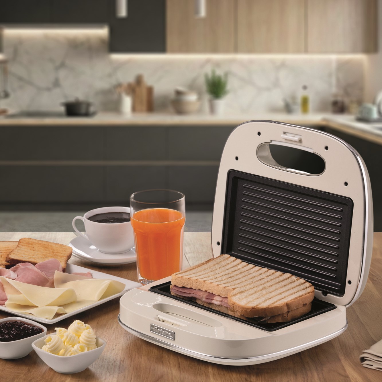 Сандвич, приготвен в тостер за сандвичи „Vintage“ с мощност 750 W, стои на кухненския плот редом с чаша портокалов сок, чаша кафе и чинии с шунка, сирене, масло и конфитюр. Наблизо е поставен елегантен електрически миксер, който идеално допълва модерната кухня на заден план.